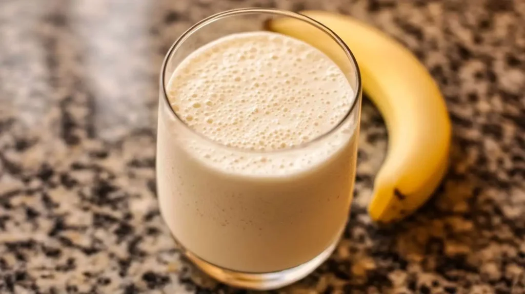 A creamy protein milkshake in a glass, placed on a polished granite countertop with a ripe banana in the background.